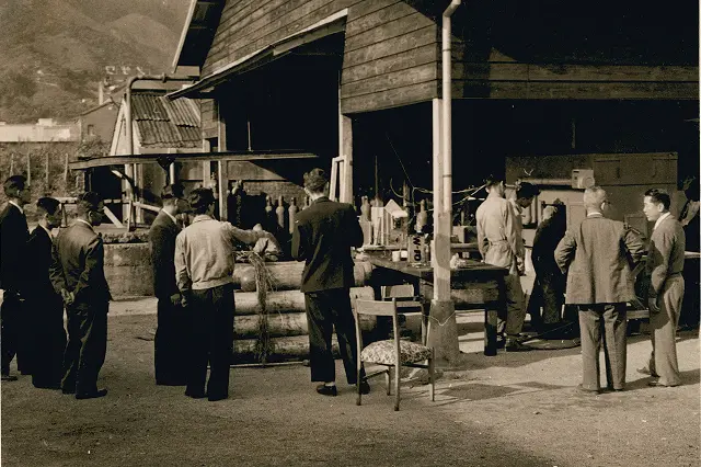 水車発電機用単一配管方式の消火装置の開発または納入前の様子。屋外に面した作業場に装置の一部（ボンベや配管）が置かれ、スーツや作業服を着た複数の関係者（技術者・顧客など）がそれを取り囲んで説明を受けている。