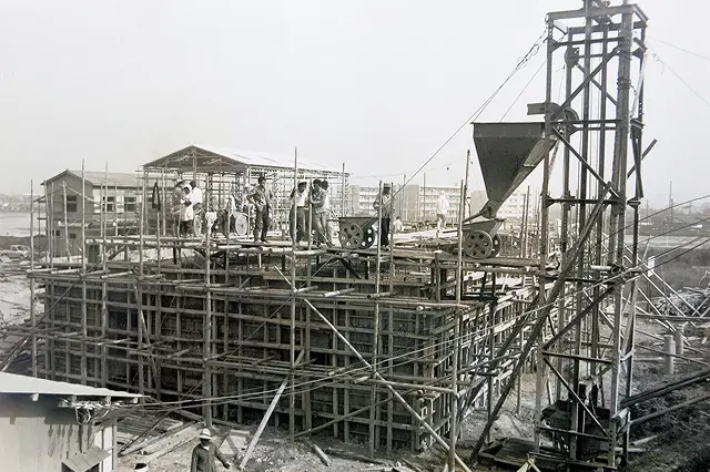 新幹線関連施設の建設現場の様子。木材の足場と型枠が組まれた基礎部分の上に、複数の作業員が立ち、建設機械（タワーとホッパー）が稼働している写真。