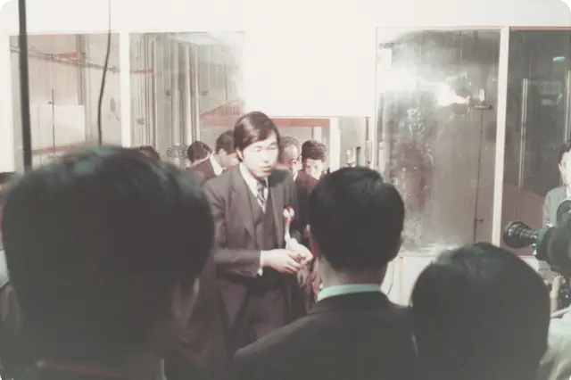 スーツ姿の担当者が中央で説明している様子を、多くの参加者や報道関係者（カメラが見える）が囲んでいる。