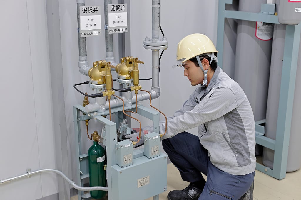 作業員が、配管やバルブが複雑に配置された消火設備を点検している。しゃがんで機器の状態を詳しく見ている。