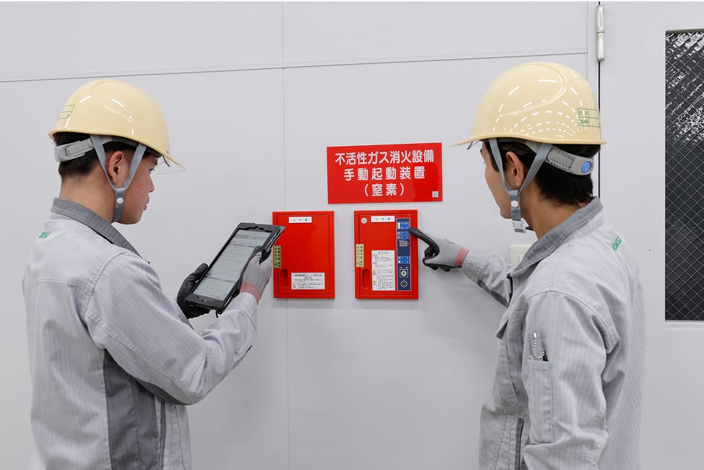 作業員2名が、不活性ガス消火設備の手動起動装置を点検している。左の作業員はタブレット端末を操作し、右の作業員は起動装置のスイッチに触れている。