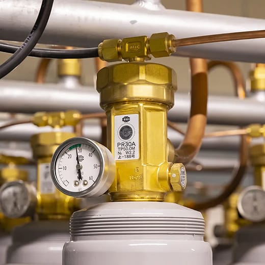 黄銅製のバルブが取り付けられた白い高圧ガス容器。バルブには圧力計、配管、型番「PR30A」のラベルが付いている